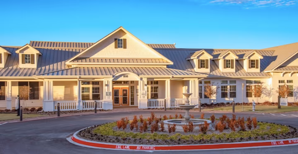 Front exterior of a light-colored senior living facility with a covered entrance, dormer windows, and a circular driveway featuring a central fountain.