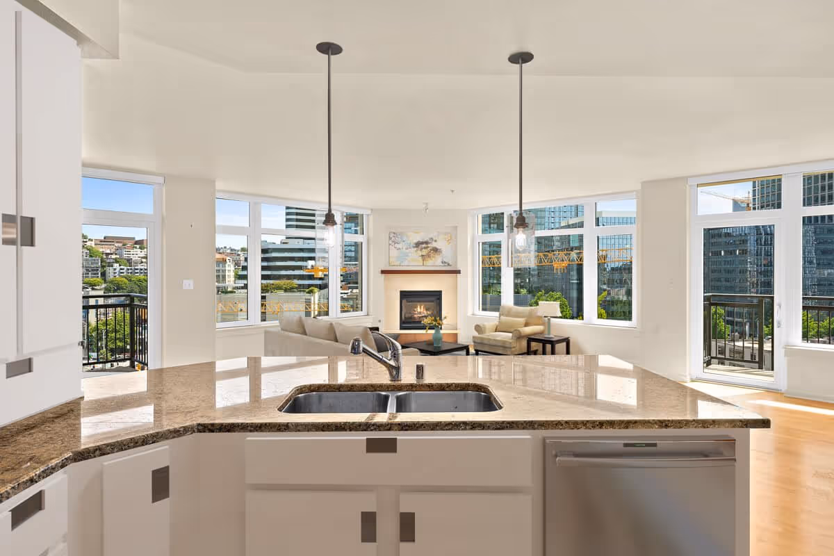 Open-concept living area with a kitchen island and sink in the foreground, seating and a fireplace by large windows overlooking the city.