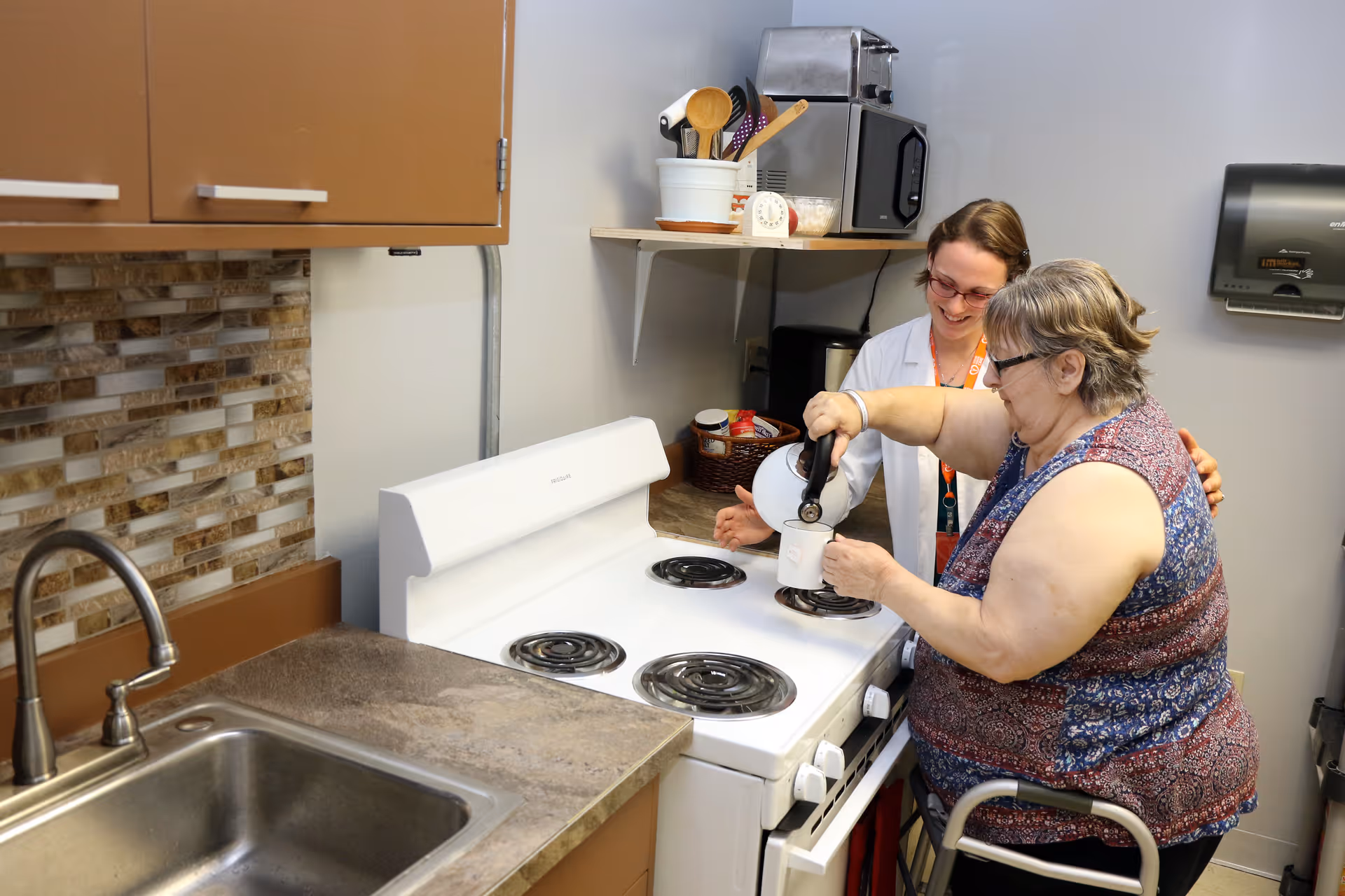 An elderly woman using a walker is pouring a beverage from a kettle into a cup on a stove, assisted by a smiling caregiver in a white coat in a kitchen setting.