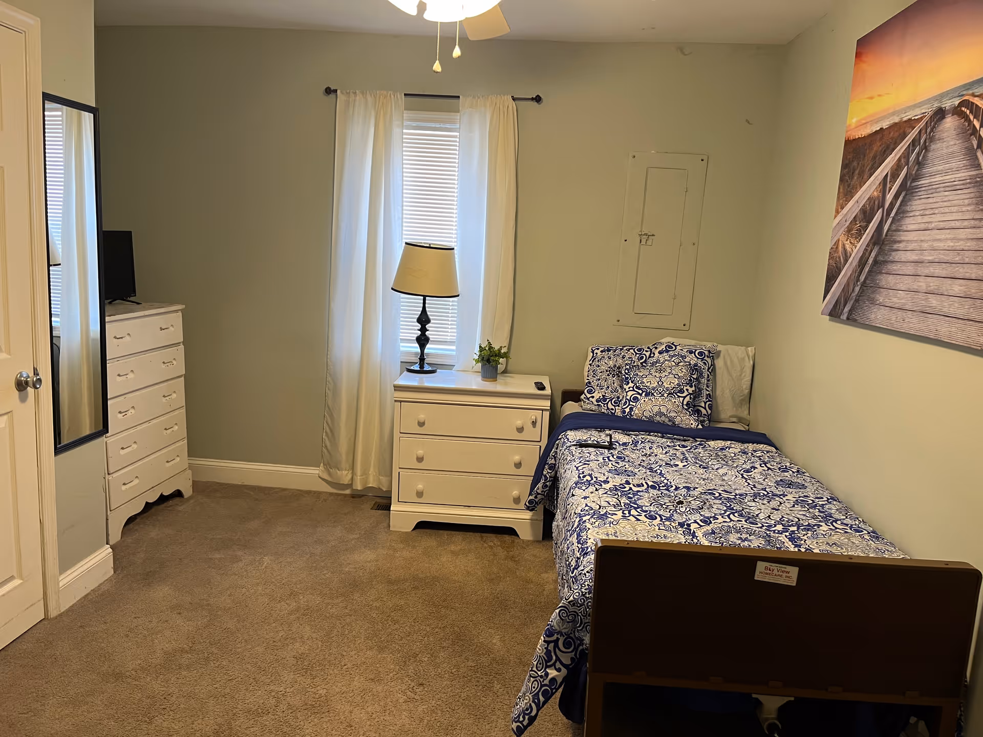 A small bedroom with a single bed covered in blue and white patterned bedding. Next to the bed is a white nightstand with a lamp and a small plant. There is a window with white curtains behind the nightstand. On the left side of the room, there is a white dresser with a TV on top and a full-length mirror mounted on the wall. The walls are painted light green, and there is a large framed picture of a boardwalk at sunset hanging above the bed.