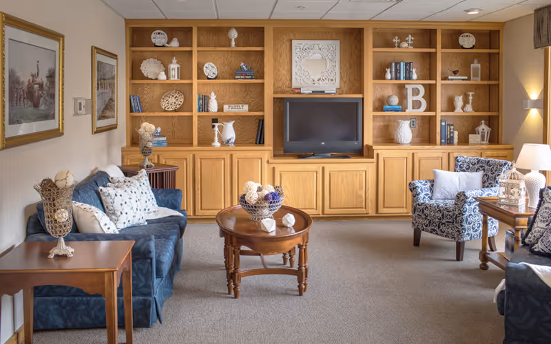 Cozy sitting room with blue upholstered sofas and armchairs, a central wooden coffee table, and built-in wooden shelving with a TV along the back wall.