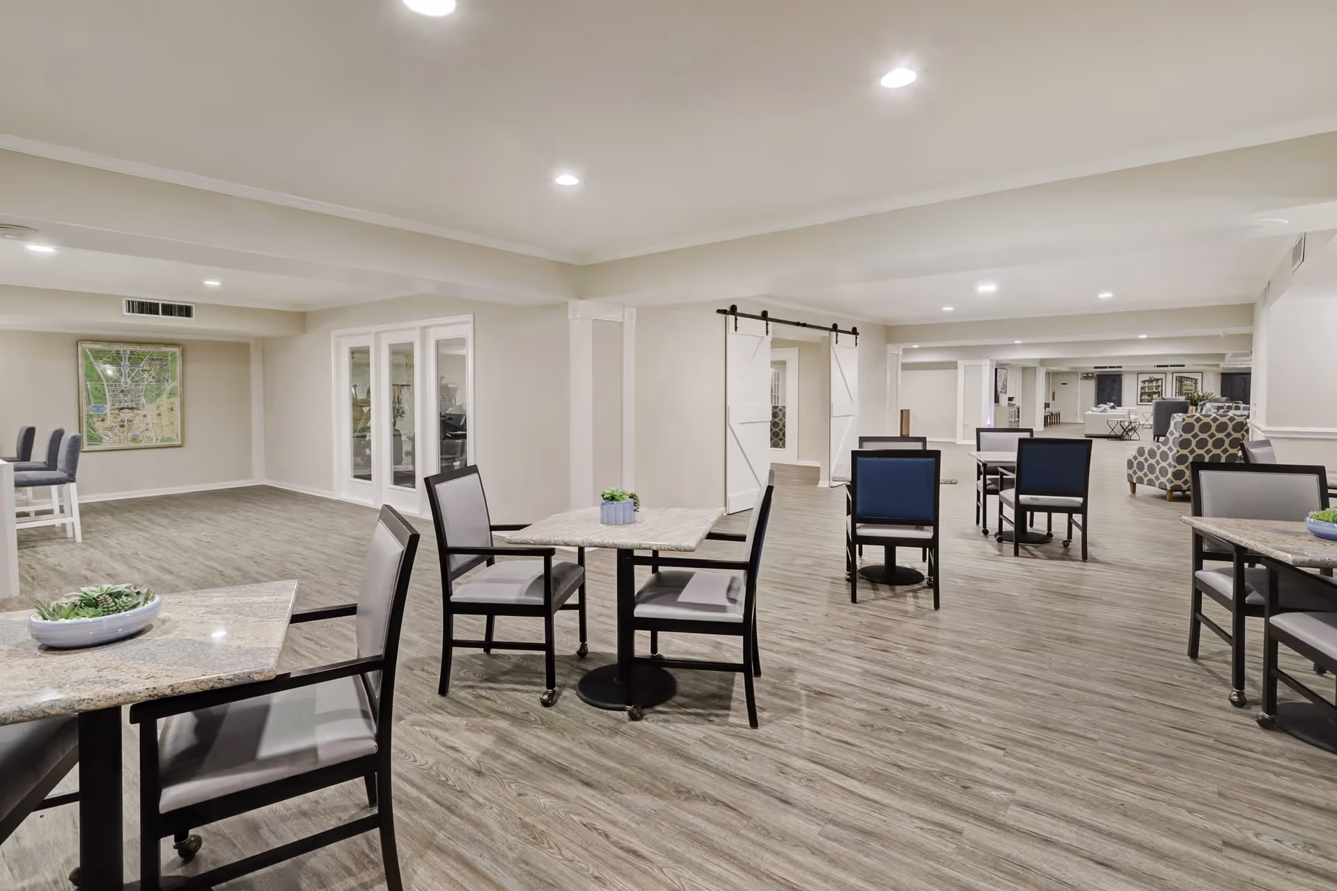 Open communal dining area with multiple tables and chairs, light wood flooring, and recessed ceiling lights.