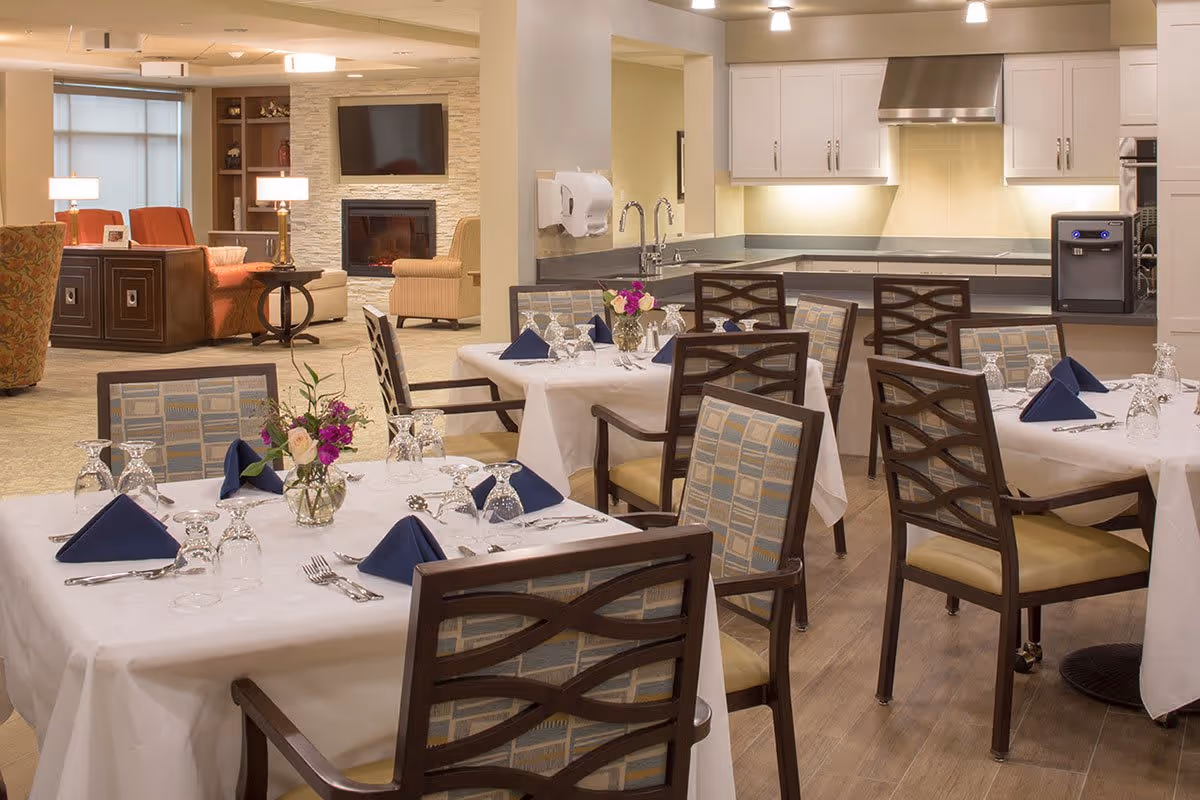 A dining area in a senior living facility with tables covered in white tablecloths, set with glassware, silverware, and navy blue napkins. Each table has a small vase with flowers. In the background, there is a kitchen area with white cabinets and a stainless steel range hood, and a living room area with armchairs, a fireplace, and a wall-mounted TV.