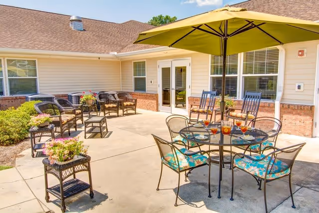 Outdoor patio area at a senior living facility with a round glass table, four chairs with floral cushions, a large green umbrella, several glasses of iced tea on the table, additional wicker chairs with cushions arranged along the building wall, and potted plants on small tables.