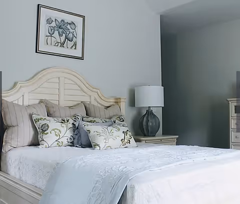 A neatly made bed with a white quilt and multiple decorative pillows in a bedroom. There is a bedside table with a lamp and a framed floral artwork hanging on the wall above the headboard.
