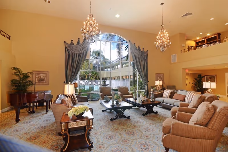 A spacious and elegant living room with large arched windows showing an outdoor view with palm trees. The room features comfortable beige and brown sofas and armchairs arranged around two dark wooden coffee tables. There are two ornate chandeliers hanging from the ceiling, a grand piano in the corner, decorative lamps on side tables, and a patterned carpet covering the floor.