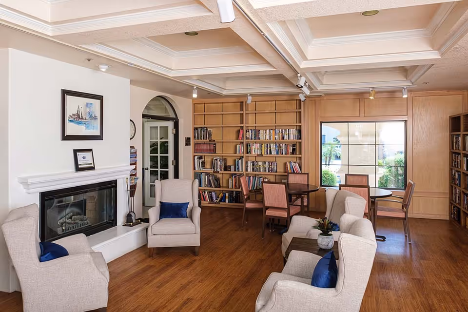 Comfortable common room with bookshelves, a fireplace, upholstered armchairs, and small tables.