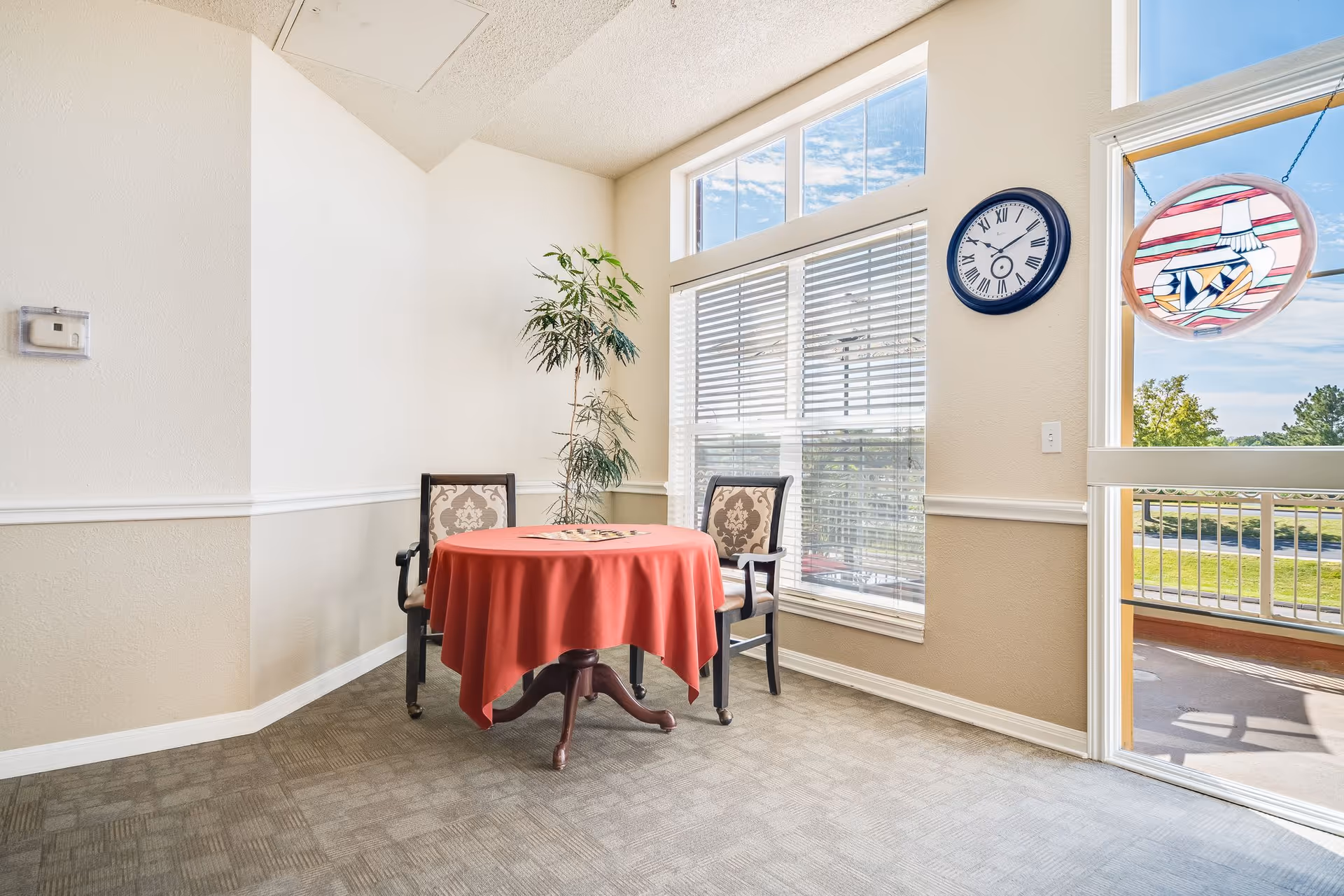 A small corner seating area with a round table covered by a red tablecloth and two upholstered chairs. There is a tall potted plant behind the chairs. Large windows with blinds allow natural light to fill the room, and a clock with Roman numerals hangs on the wall next to a glass door leading to an outdoor balcony with a stained glass decoration hanging on the door.