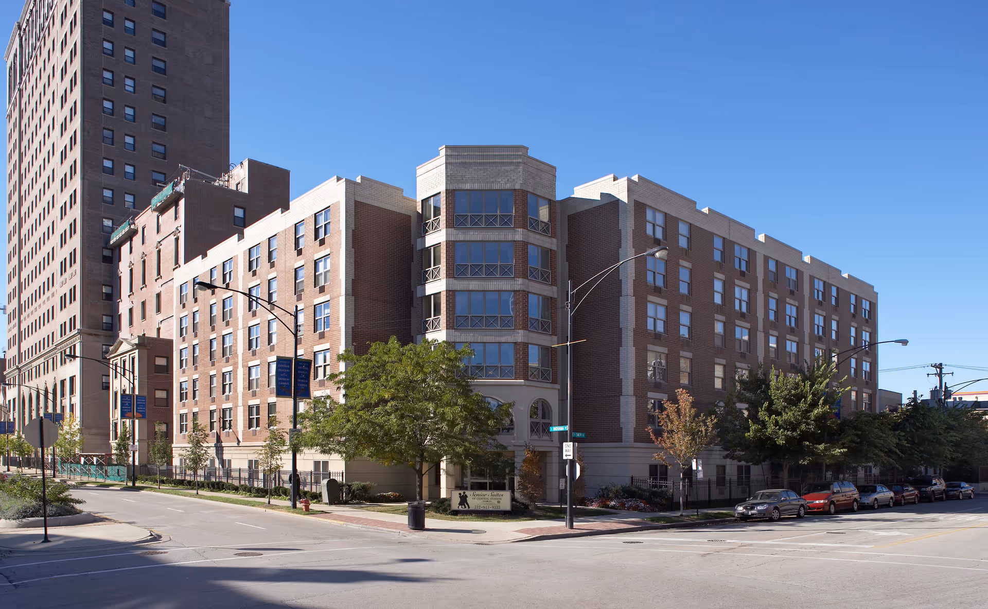 Exterior view of a multi-story brick building named Senior Suites of Central Station, located at a street corner with trees and parked cars along the street under a clear blue sky.
