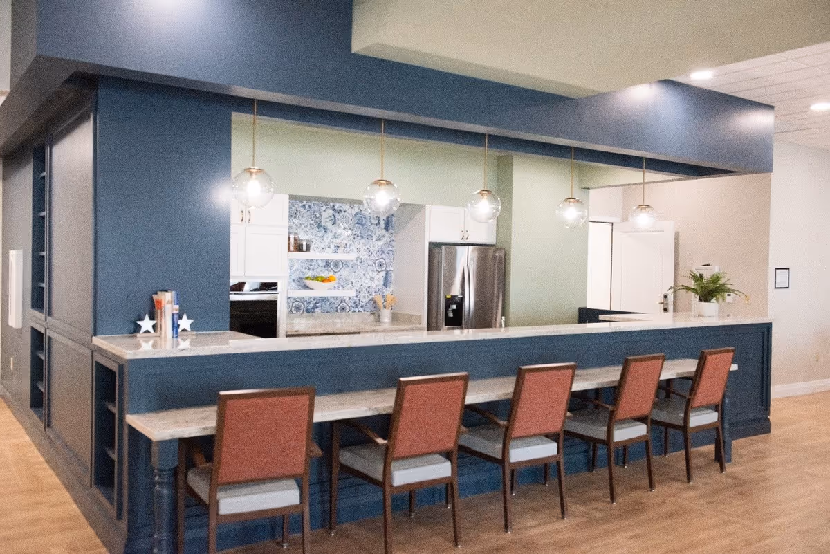 A modern kitchen area with a long marble countertop bar and six chairs with reddish-brown backs and gray seats. The kitchen has blue cabinetry, pendant lights hanging above the bar, a stainless steel refrigerator, and a decorative blue and white tile backsplash. The floor is wooden, and there is a small plant on the right side of the counter.