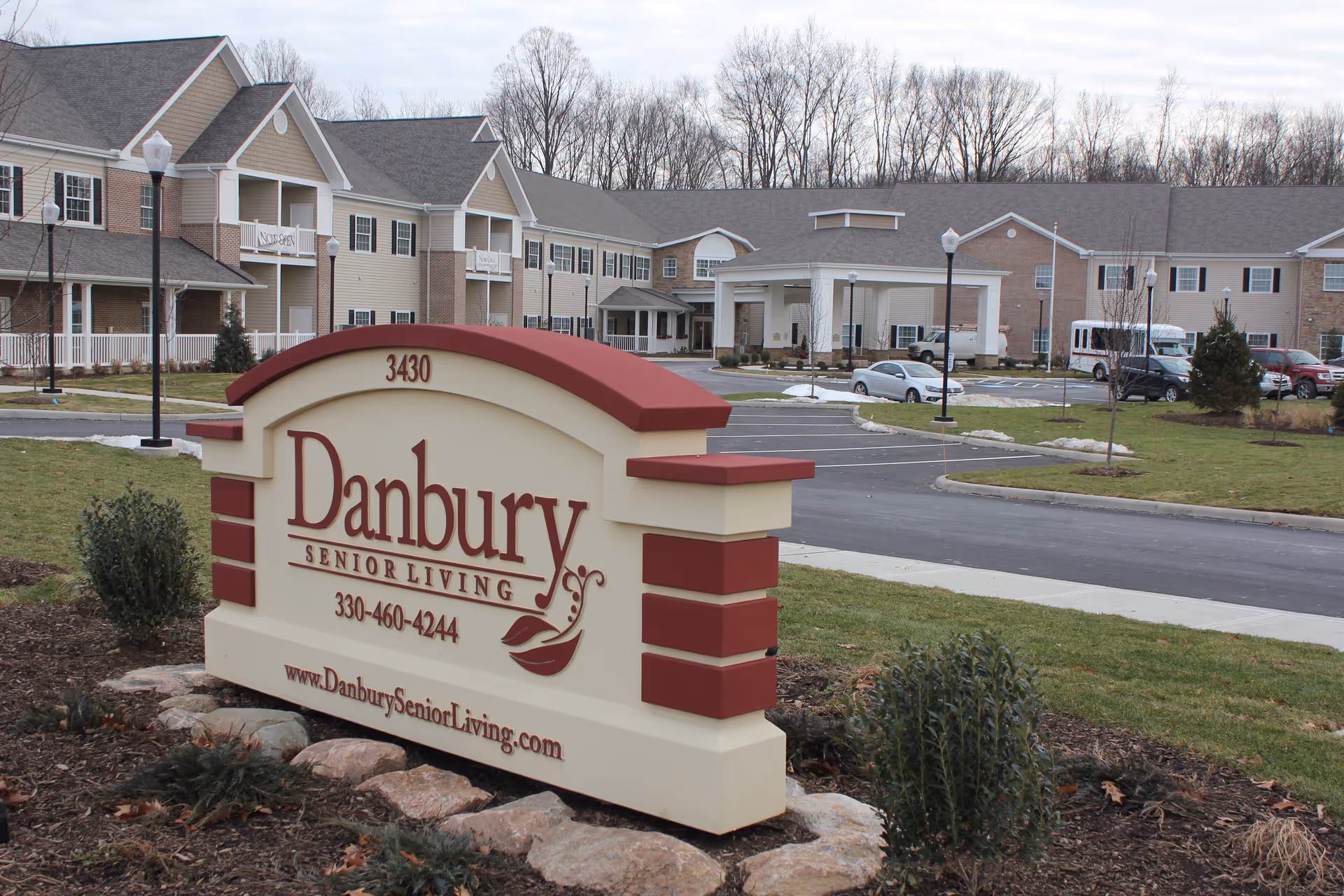 Exterior view of Danbury Senior Living facility showing a large sign with the name, phone number, and website in front of a two-story building with multiple windows, balconies, and a covered entrance. The surrounding area includes a parking lot, street lamps, some cars, and landscaped greenery with trees in the background.
