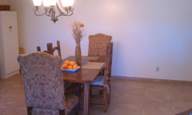 A dining room with a wooden table and four upholstered chairs. On the table, there is a vase with dried flowers and a tray with oranges. A chandelier hangs above the table, and there is a white cabinet against the wall in the background.
