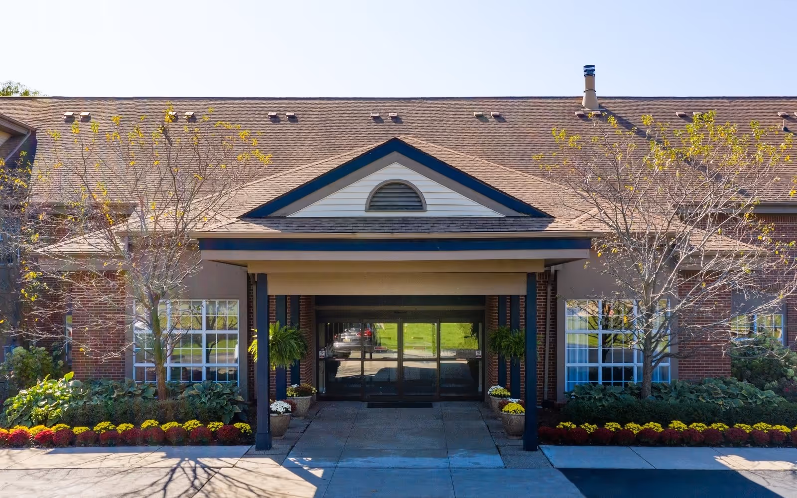 Front exterior view of a senior living facility named Brookhaven Manor with a covered entrance, brick walls, large windows, and landscaped flower beds with small trees on either side.