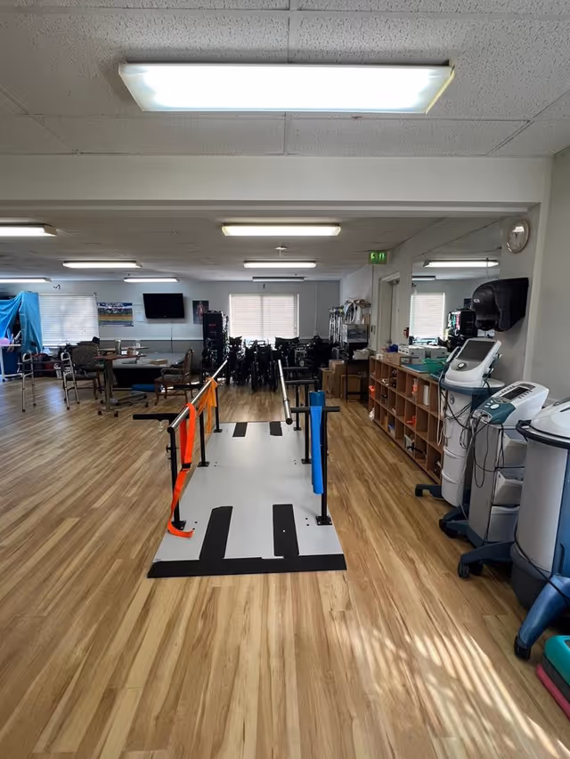 Interior of a rehabilitation or therapy room with wooden flooring, parallel bars for walking exercises in the center, various medical and therapy equipment on the right side, chairs and tables on the left, and wheelchairs lined up at the back near windows with blinds.