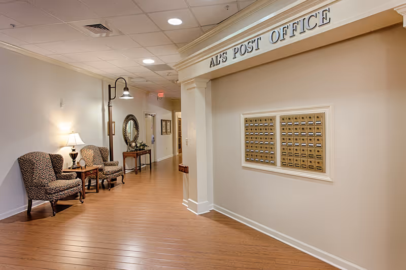 Interior hallway of an assisted living facility with a section labeled 'Al's Post Office' featuring multiple mailboxes mounted on the wall. The hallway has wooden flooring, two patterned armchairs with a small table and lamp between them, a tall floor lamp, a decorative mirror, and a small table with flowers further down the hall.