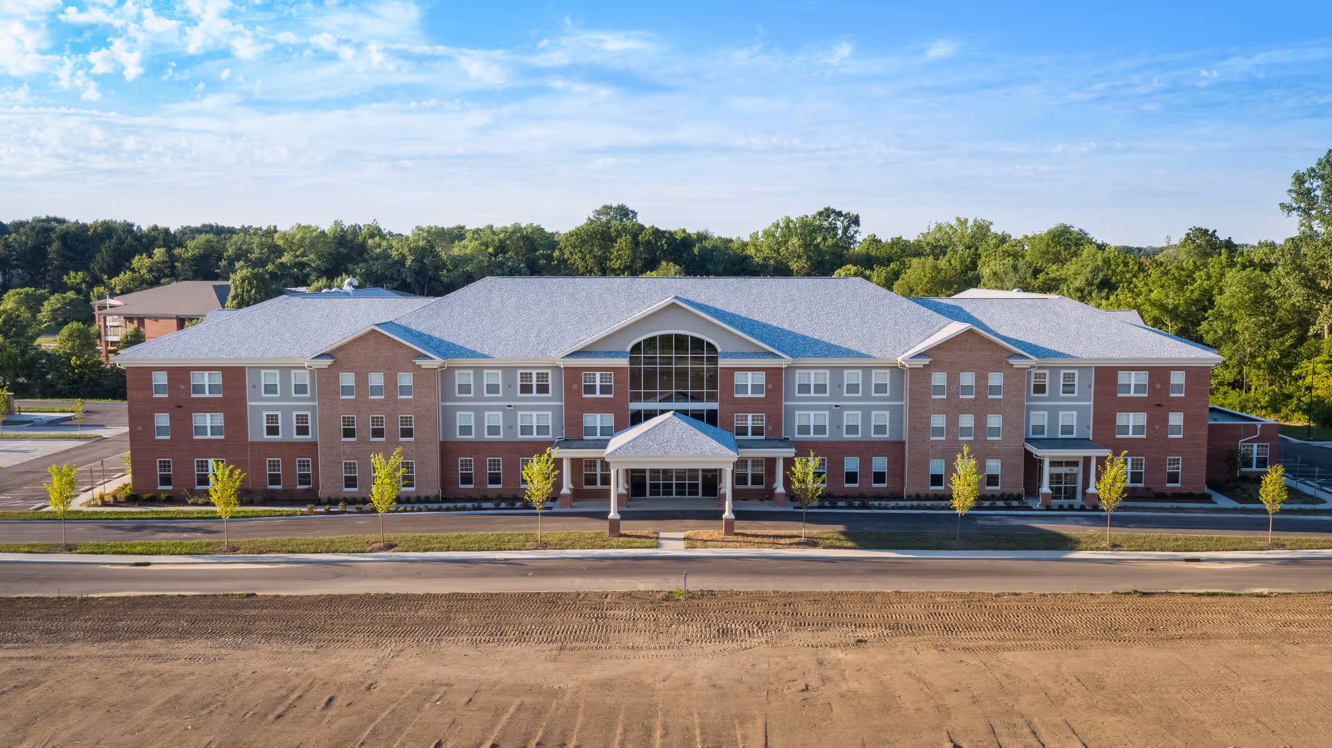 Front exterior view of a large, three-story senior living facility building with a combination of red brick and gray siding, multiple windows, a central entrance with a covered porch, and surrounded by young trees and greenery under a partly cloudy sky.