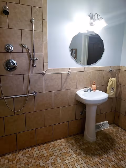 A bathroom with beige tiled walls and floor, featuring a shower with a handheld showerhead and grab bar, a white pedestal sink with a soap dispenser, a towel ring holding a yellow towel, a decorative mirror above the sink, and a wall-mounted light fixture.