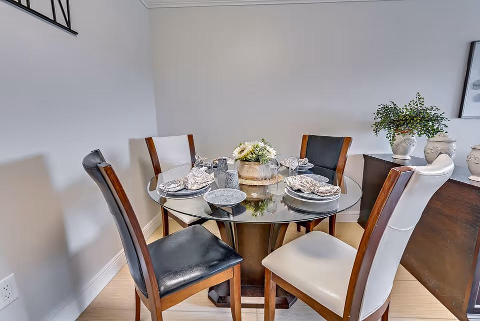 A small dining area with a round glass table set for four people. The table has four chairs with wooden frames and black and white cushioned seats. The table is set with plates, napkins, and glasses, and there is a floral centerpiece. In the background, there is a dark wooden sideboard with decorative vases and a green plant.