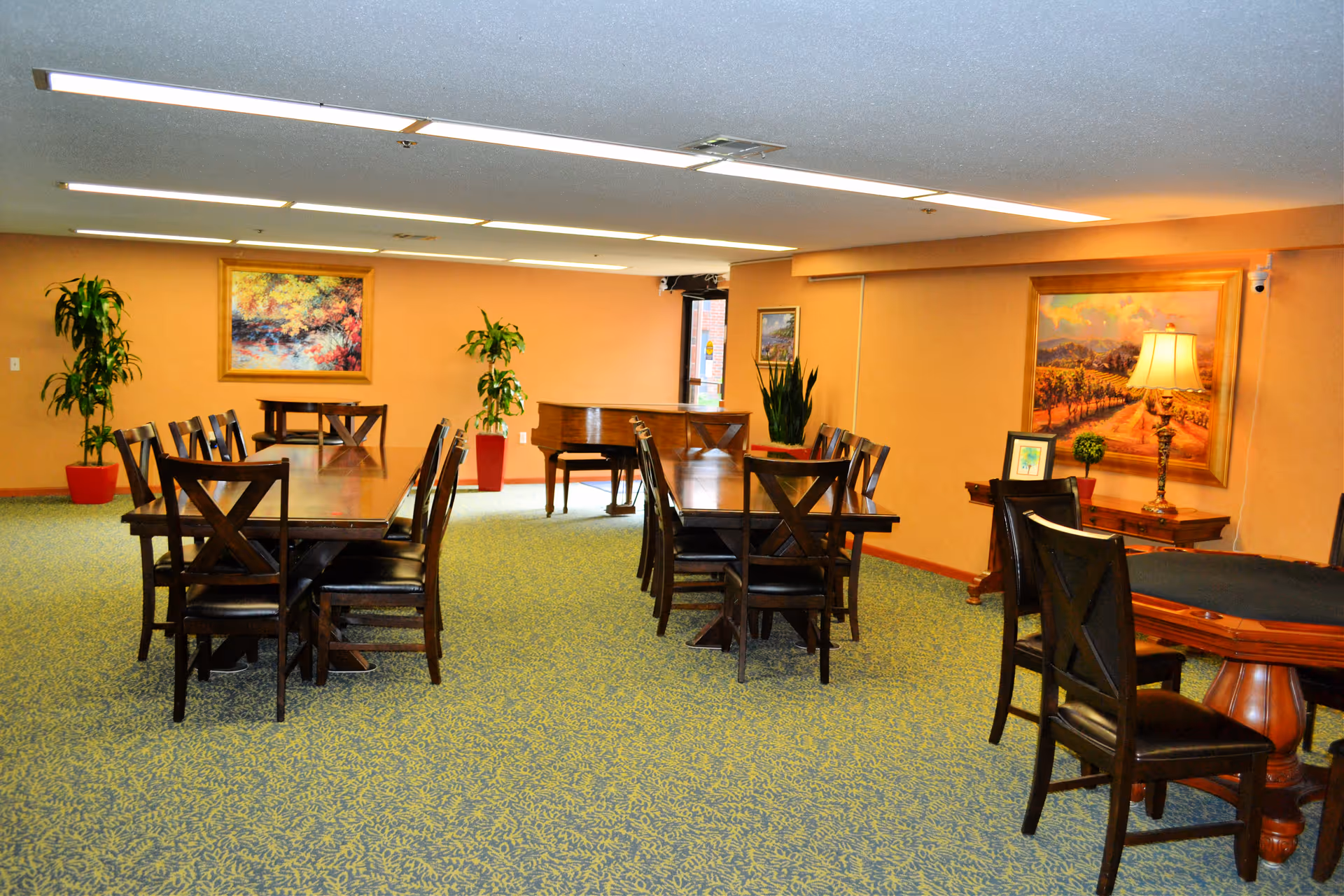 A brightly lit common dining/activity room with several wooden tables and chairs, potted plants, and framed paintings on the walls.