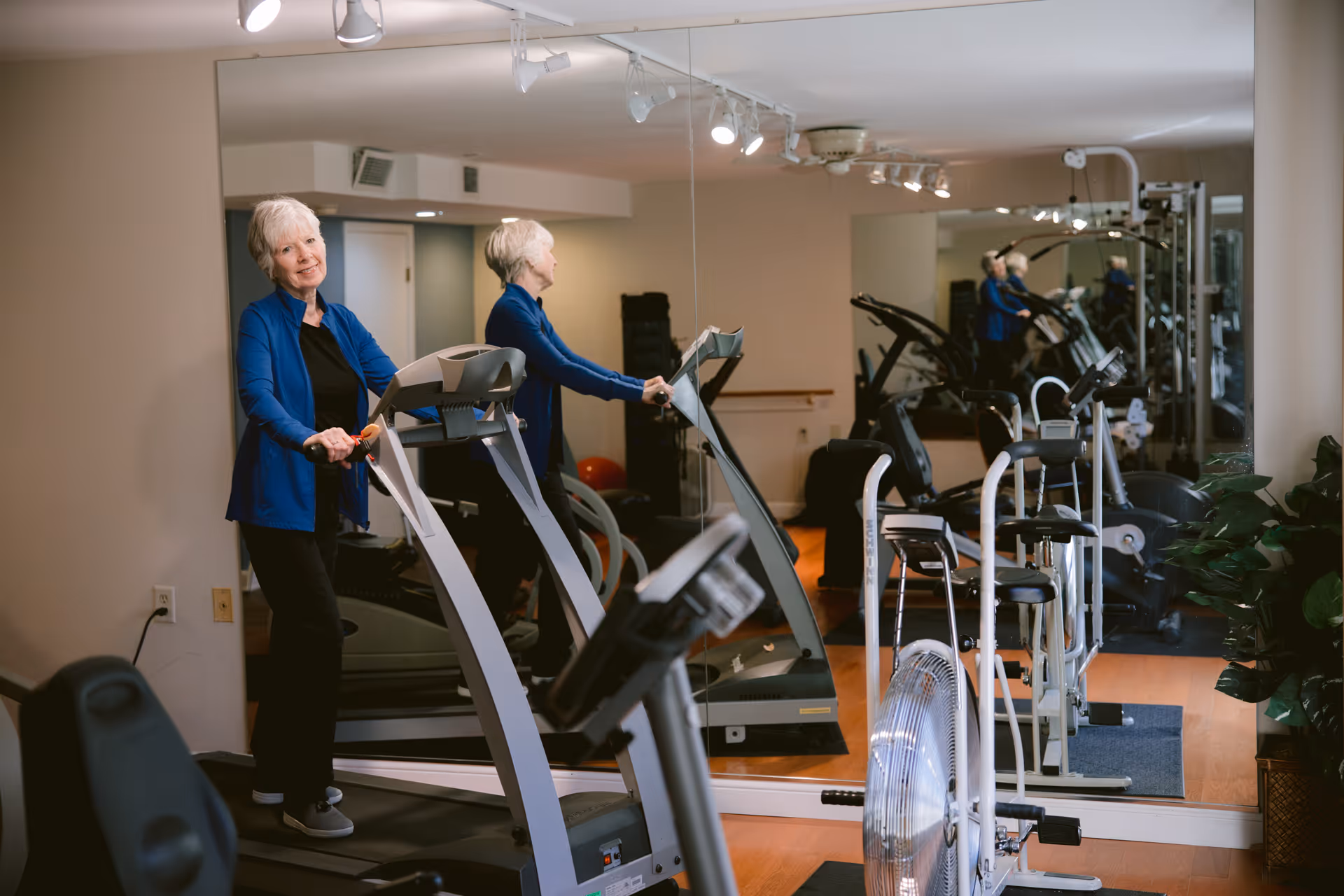 An elderly woman in a blue jacket and black pants standing on a treadmill in a fitness room with exercise equipment and a large mirror on the wall.