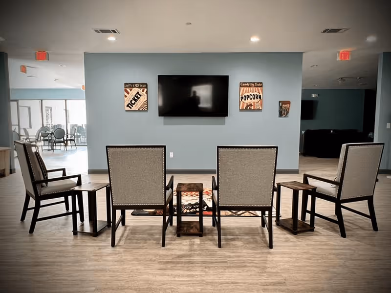 Row of four armchairs with side tables facing a wall-mounted TV on a blue wall in a communal lounge.