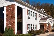 Exterior view of a two-story senior living facility building with white siding and red brick accents, multiple windows, and a small covered entrance. There are shrubs and greenery along the building and trees in the background under a clear sky.