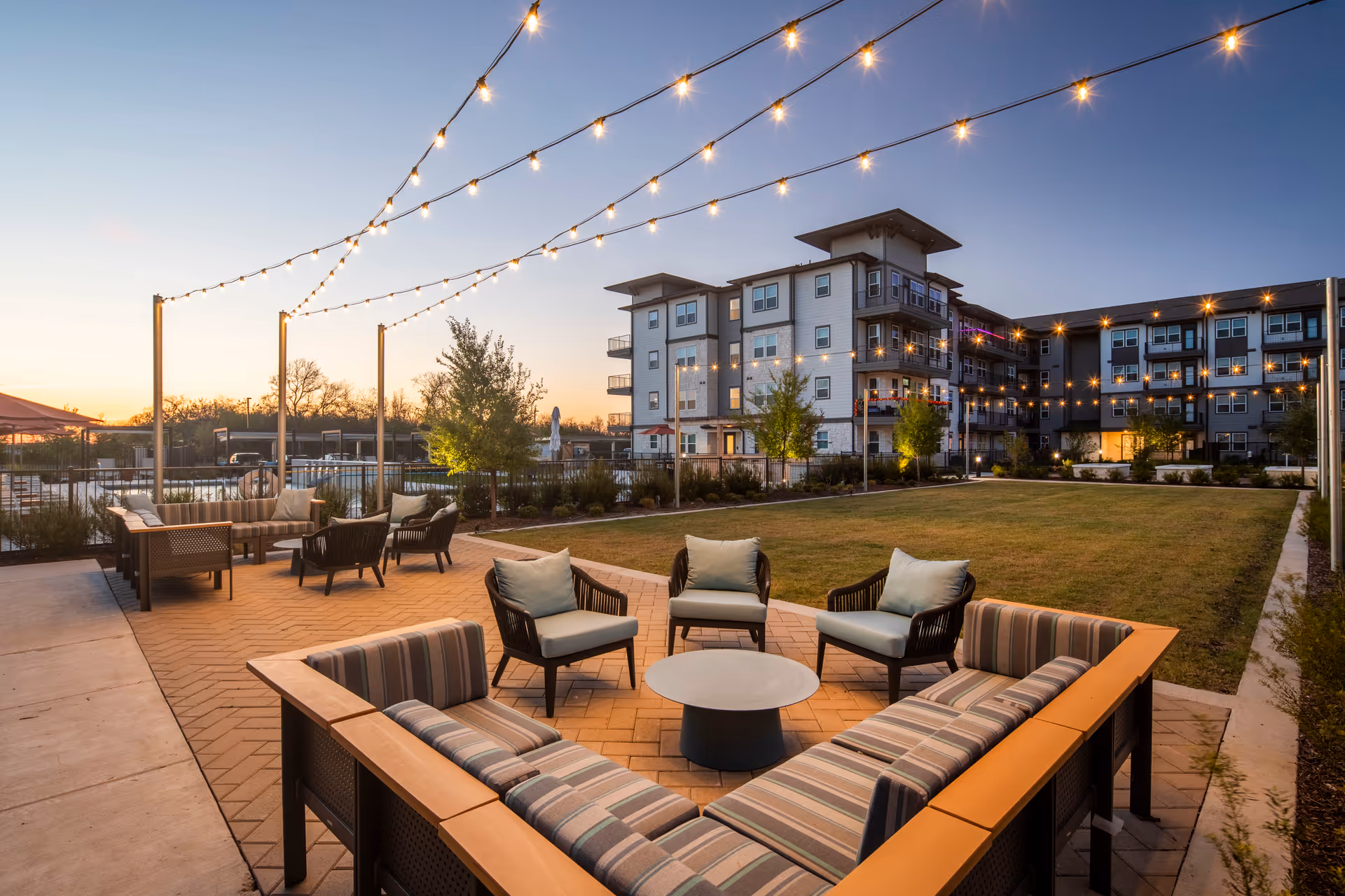 Outdoor courtyard at dusk with string lights, patio seating around a low table, a grassy lawn, and a multi-story residential building in the background.
