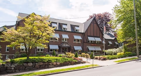 Exterior view of a large brick building with multiple windows, some with white awnings, surrounded by trees and landscaping. The building has a steep roof with dormer windows and a sign near the entrance that reads 'Southmayd Home'.