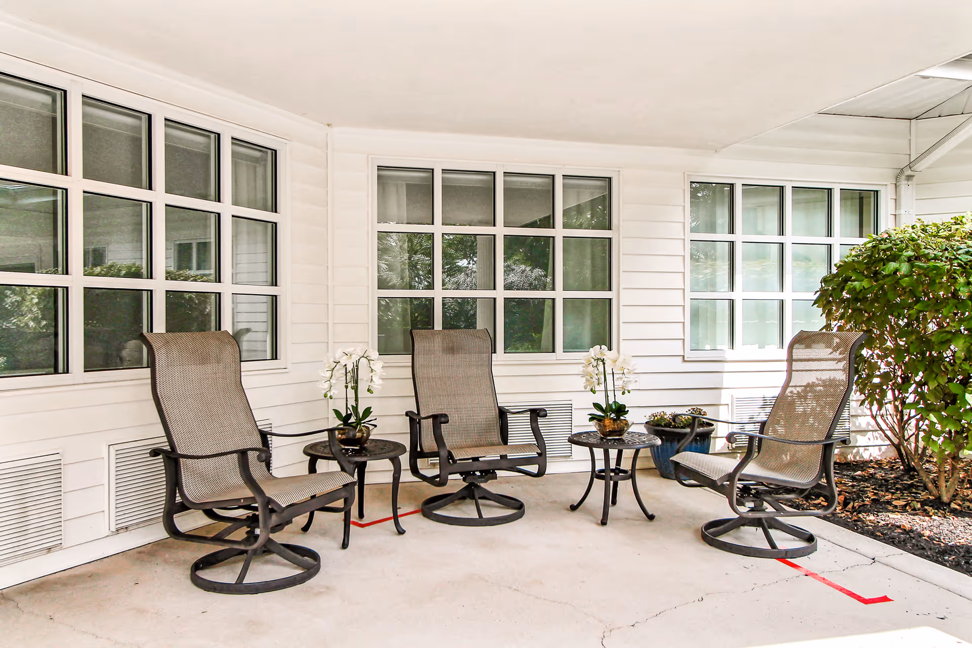 Covered outdoor patio with four swivel mesh chairs arranged around small tables and potted plants in front of white siding and windows.