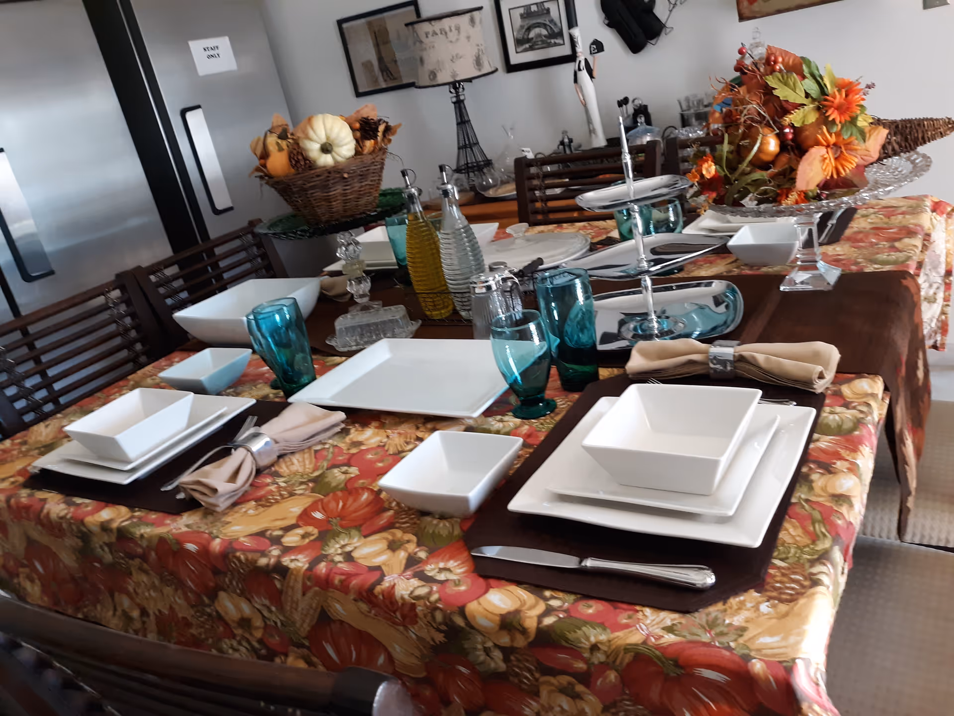 A dining table set for a meal with white square plates, bowls, beige napkins with silver napkin rings, blue drinking glasses, and silverware. The table is covered with a colorful tablecloth featuring autumn-themed patterns. There are decorative centerpieces including a basket with small pumpkins and a glass dish with fall flowers and leaves. In the background, there are chairs, a lamp with an Eiffel Tower design, and a door labeled 'Staff Only'.