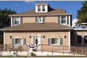 Two-story beige assisted living building with front porch, bunting, and surrounding greenery.