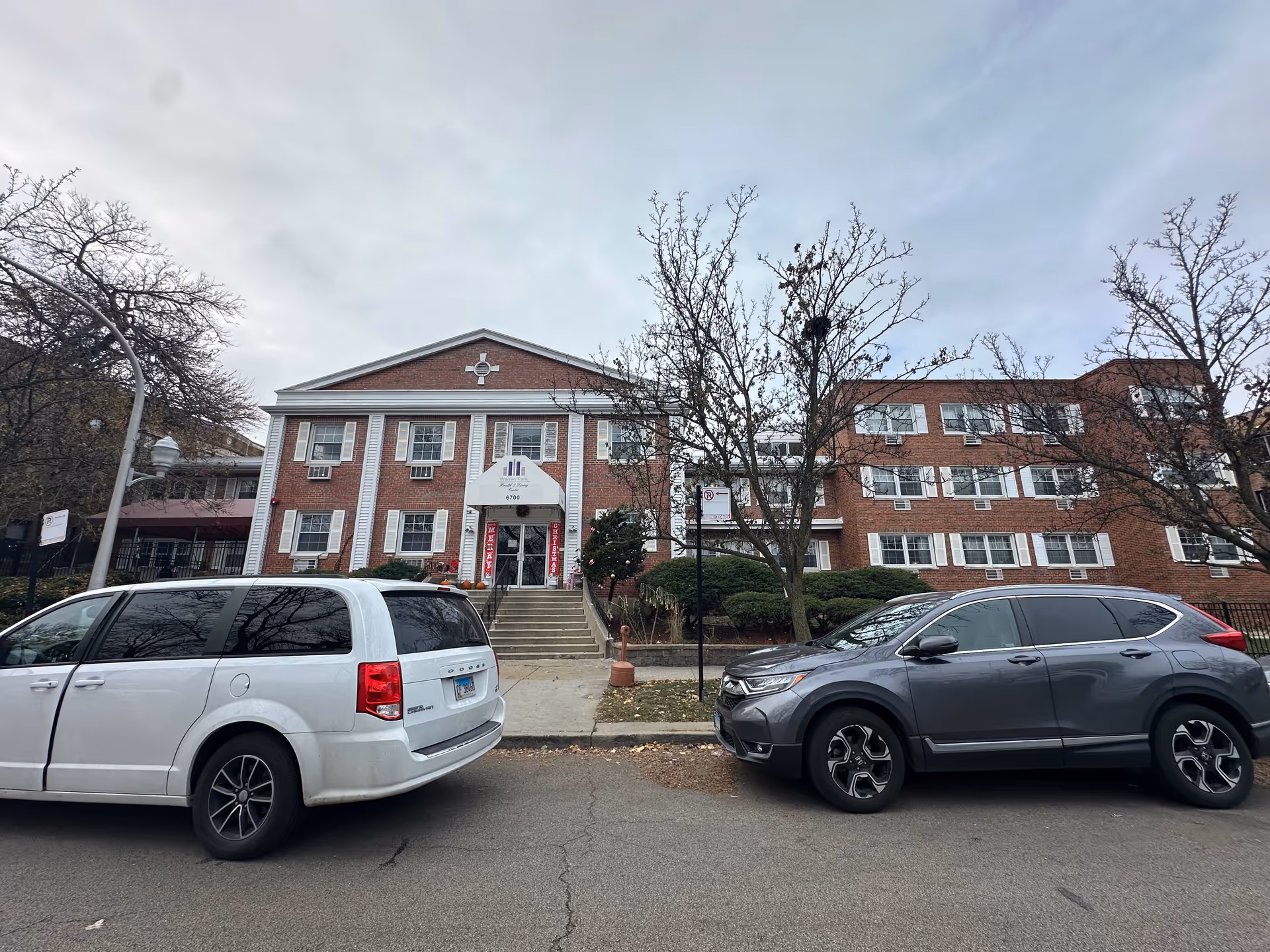Front exterior view of a three-story brick building with white window shutters and a central entrance with stairs leading up to it. Two cars are parked on the street in front of the building. Leafless trees and some bushes are visible around the entrance area under a cloudy sky.