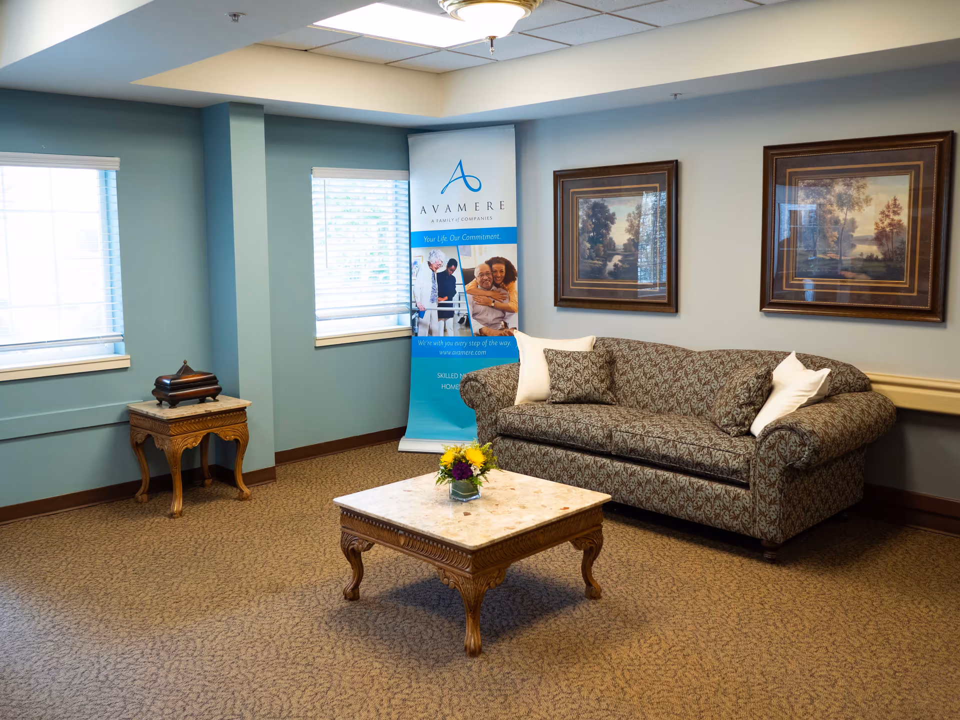 A cozy sitting area in a senior living facility with a patterned sofa adorned with three pillows, a wooden coffee table with a small flower arrangement, two framed landscape paintings on the wall, two windows with blinds, and a promotional banner for Avamere in the corner.