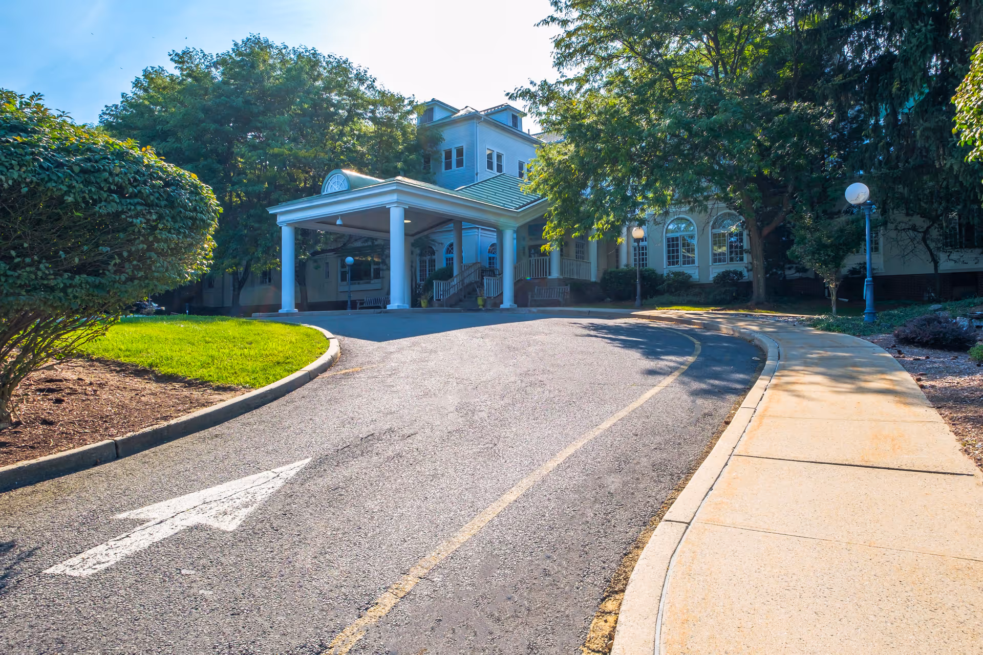 Entrance driveway to a senior living facility with a covered drop-off area supported by white columns, surrounded by green trees and bushes under a clear blue sky.