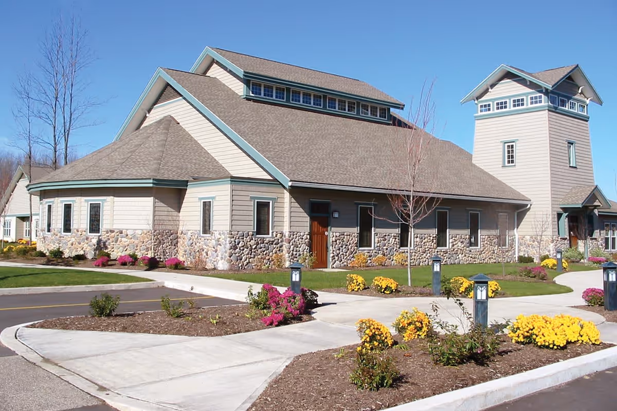 Front exterior of the Cherry Ridge senior living building with stone-and-siding facade, a tower, landscaped flower beds and paved walkways under a clear blue sky.