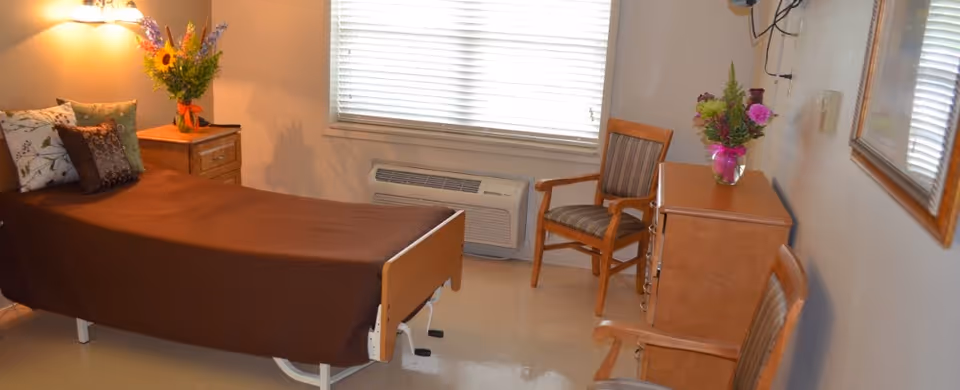 A nursing facility bedroom with a single bed covered in a brown blanket and several decorative pillows. There is a wooden nightstand with a lamp and a vase of flowers next to the bed. Across from the bed, there is a wooden dresser with a vase of flowers on top and two wooden chairs with striped cushions. A window with blinds and an air conditioning unit is on the wall behind the dresser.