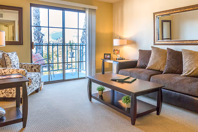 A bright living room with a large brown sofa adorned with multiple cushions, a patterned armchair, a dark wooden coffee table with two small potted plants underneath, and a side table with a lamp and framed photo. The room features a large window with sliding glass doors leading to a balcony with outdoor seating and a view of palm trees and mountains in the background.