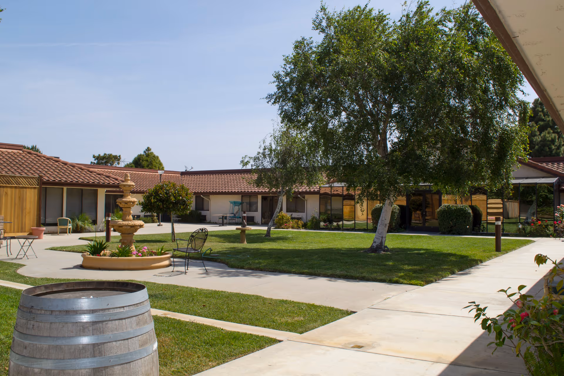 Outdoor courtyard area of a senior living facility with a large tree, a multi-tiered fountain surrounded by flowers, metal chairs, and a wooden barrel. The courtyard is surrounded by single-story buildings with tiled roofs and large windows.
