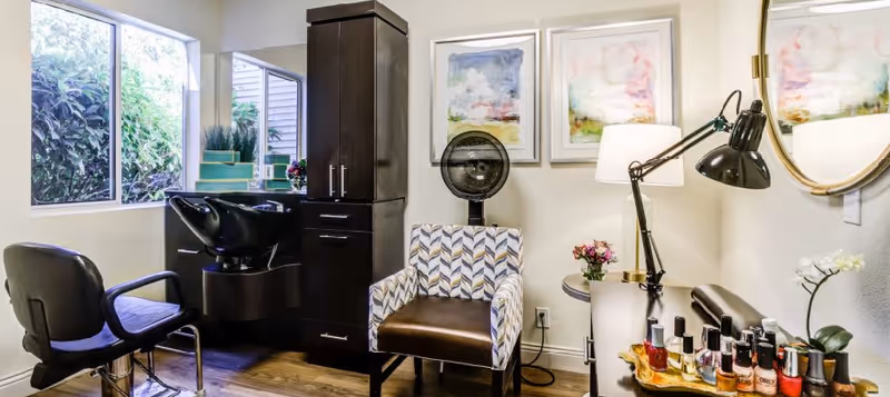 A sunlit salon room featuring a hair-wash sink and styling chairs, a patterned armchair with a hooded dryer, and a table displaying nail polishes and lamps.