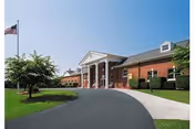 Exterior view of a single-story brick building with white columns at the entrance, a curved driveway, green lawn, trees, and an American flag on a flagpole against a clear blue sky.
