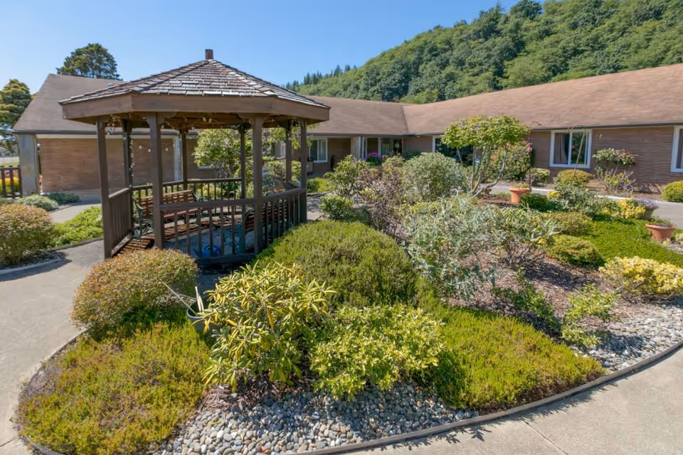 A landscaped outdoor garden area with various shrubs and plants surrounding a wooden gazebo with a shingled roof. The garden is bordered by a curved concrete walkway, and a single-story building with windows and a brown roof is visible in the background, set against a backdrop of green trees and a clear blue sky.