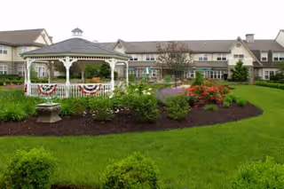 A large senior living facility building with beige walls and a brown roof, viewed from a landscaped garden area. The garden features a white gazebo decorated with red, white, and blue bunting, surrounded by green grass, bushes, and colorful flowers.