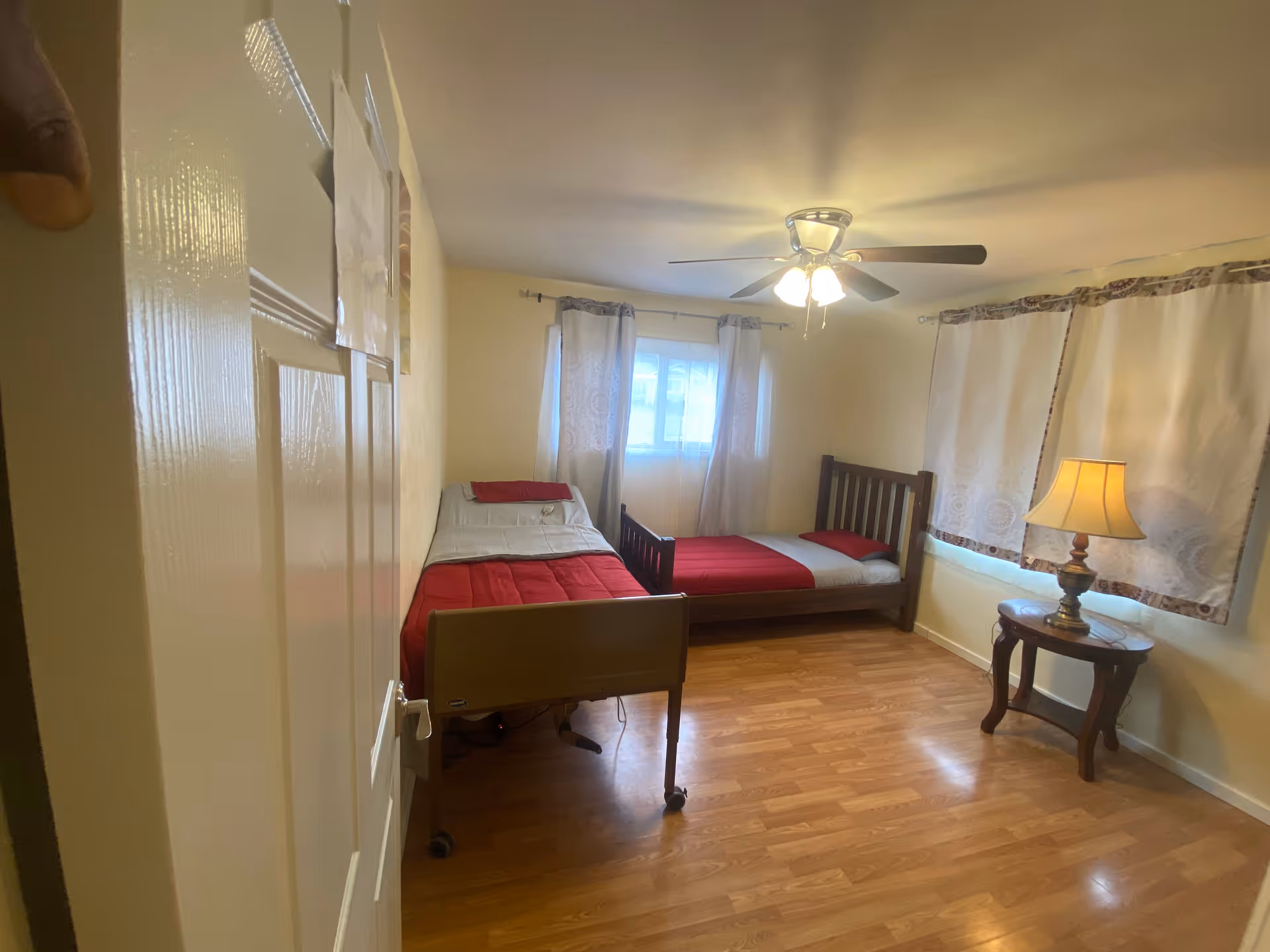 Small bedroom with two single beds, a ceiling fan, wood floor, and a side table with a lamp.
