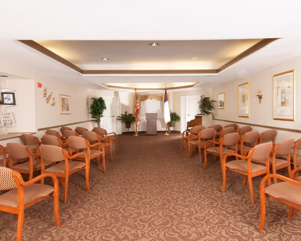 A meeting or event room with rows of wooden chairs with patterned cushions arranged facing a podium at the front. The room has a patterned carpet, framed pictures on the walls, two flags behind the podium, and several potted plants.