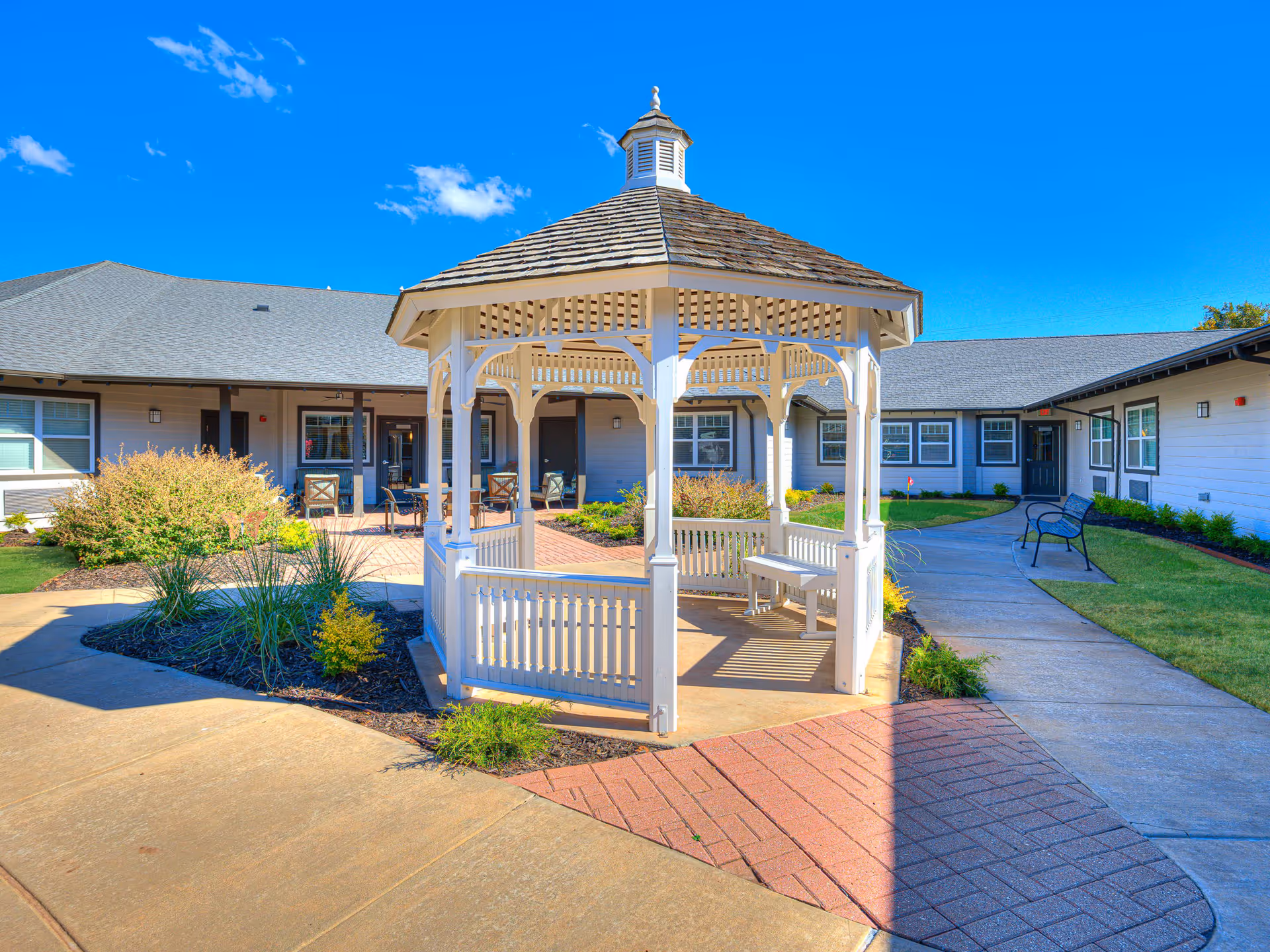 A bright outdoor courtyard area at Iris Memory Care of Nichols Hills featuring a white wooden gazebo with a bench inside, surrounded by paved walkways, green grass, shrubs, and a building with multiple windows and doors in the background under a clear blue sky.