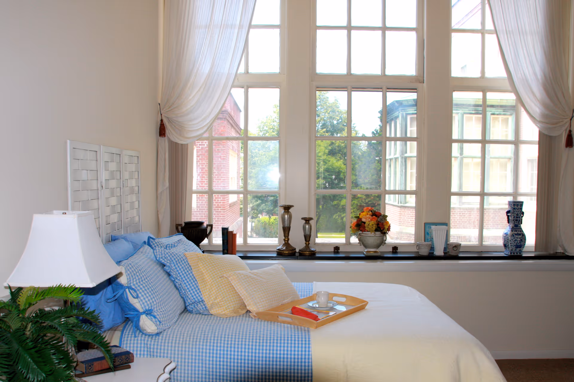 A bright bedroom with a large window letting in natural light. The bed is made with blue and white checkered bedding and several pillows. A wooden tray with a cup and a red napkin rests on the bed. On the windowsill are decorative items including candlesticks, a flower vase with colorful flowers, books, and a blue and white ceramic vase. White curtains frame the window, and a white bedside lamp and green plant are visible on a small table next to the bed.