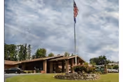 Exterior view of a single-story brick building with a sloped roof, an American flag flying on a flagpole in front, surrounded by green grass and trees under a partly cloudy sky.