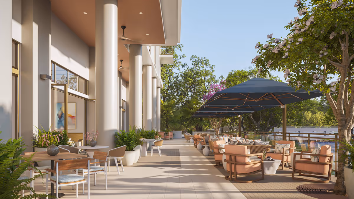 Outdoor patio area at Vi at Aventura featuring comfortable seating with cushioned chairs and sofas under large blue umbrellas, surrounded by trees and plants. The patio is adjacent to a building with large windows and tall columns, with tables and chairs arranged for relaxation and socializing.