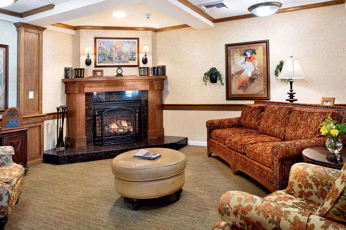 Cozy living room with a lit fireplace surrounded by a wooden mantel. The room features a patterned red sofa, a floral armchair, a round beige ottoman with a magazine on top, a side table with a vase of yellow flowers, and framed artwork on the walls. Warm lighting and beige carpeting complete the inviting atmosphere.