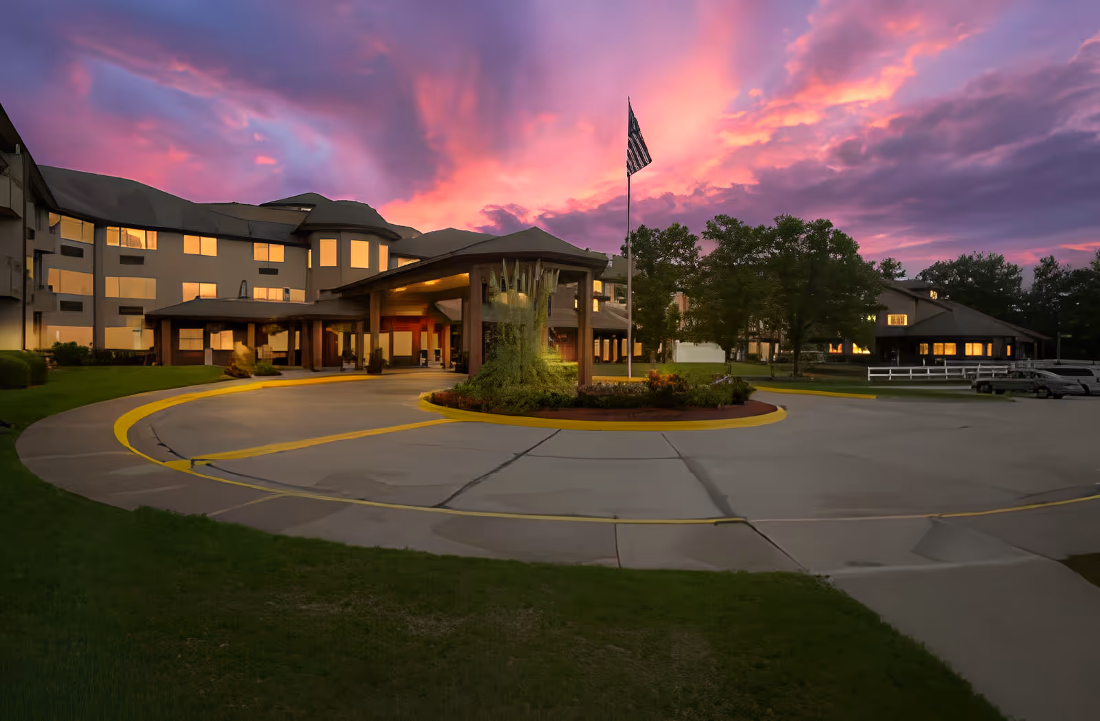 Front entrance of Holiday Country Squire with a covered circular driveway, flagpole, and a dramatic pink-purple sunset sky.